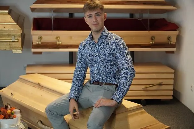 Der angehende Bestatter Luis Bauer sitzt auf einem Holz-Sarg für ein Foto.